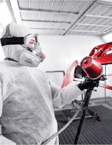 Protective worker in a cleanroom environment spray painting a red vehicle with a spray gun, wearing full safety gear including a suit, gloves, goggles, and a mask for auto painting and car refinishing.